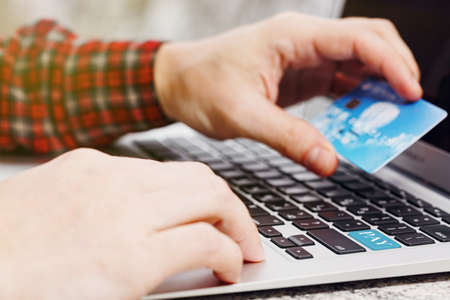 Man in checkered shirt holding a credit card for making an online purchase via laptop. Enter key on keyboard replaced with pay tag. One touch payment concept. Close-up capture, selective focus.の写真素材