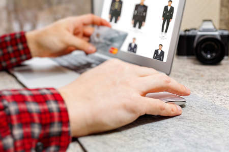 Male person in checkered shirt doing online apparel shopping and ready to make payment via credit card. Online clothes store web page opened on laptop screen. Close-up capture, selective focus.の写真素材