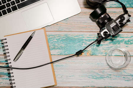 Photographer workspace - laptop, vintage DSLR camera wired to computer, notepad with pen and glass of water on vintage style rough painted wooden table. Freelancer home office concept. Top view.の写真素材