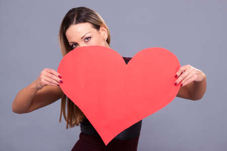Valentines Day. Woman wearing red dress holding big heart sign love symbol on grey backgroundの写真素材