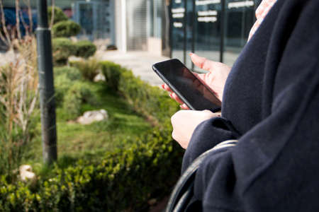 European woman with blonde hair. In a dark blue coat. Holding a black, mobile phone. Horizontal photo. Blurred background. Concept: mobile technologies, taxi call.の写真素材