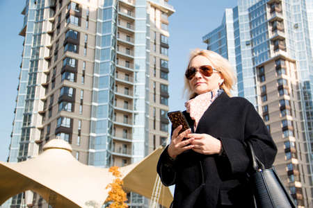 European woman with blonde hair. In a dark blue coat. Holding a black, mobile phone. Horizontal photo. Blurred background. Concept: mobile technologies, taxi callの写真素材