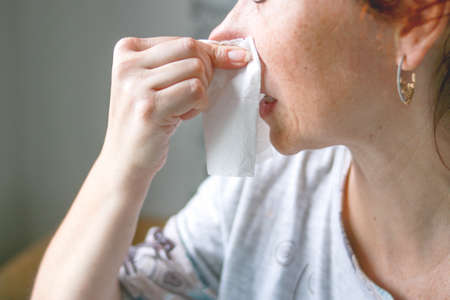 Middle-aged woman. With red hair. He blows his nose into a paper napkin. On a gray background. Horizontal photo. Concept: disease, virus, treatment, medicine, cold.の写真素材