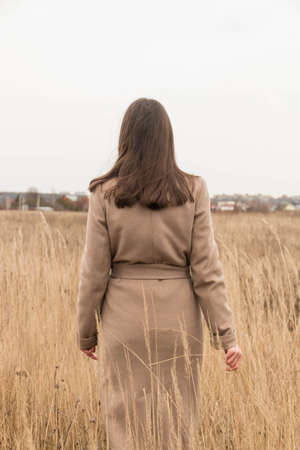 A young girl of European nationality with long hair. Brunette. In a beige, long coat. Stands with his back to a field of reeds. Spring. Autumn.Day. Cloudy weather. Vertical photoの写真素材