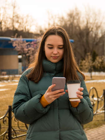 Beautiful girl, brunette with long hair, European appearance. In everyday clothes. Walking around the city with a Cup of coffee and a mobile phone in his hands. Spring. Autumn. Day. Cloudy weather.の写真素材