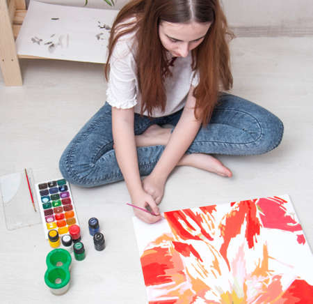 A girl of European nationality, a brunette with long hair, a teenager. Draws a picture on canvas with paints, sitting on the floor in the bedroom. In everyday clothes. The child is at home, in quarantine. The view from the top. Day.の写真素材