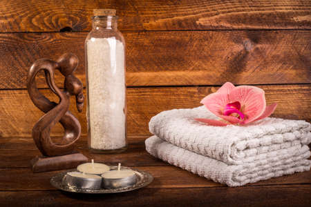 Spa or wellness set. White sea salt in white glass bottle, candle, towels, brown wooden statuette and pink flowers lily on brown wooden background. Selective focus. Place for text. Health concept. Flat lay.の写真素材