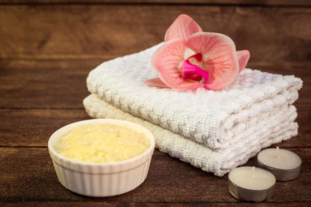 Spa or wellness set. Yellow sea salt in white bowl, towels, candle and pink flowers lily on brown wooden background. Selective focus. Place for text. Health concept. Flat lay.の写真素材