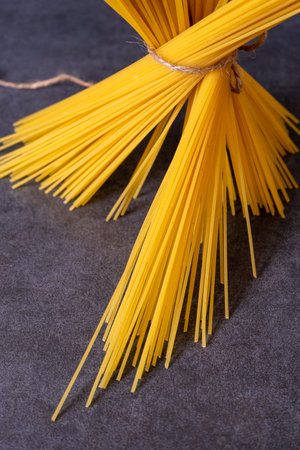 Cooking Italian pasta concept. A bunch of raw yellow spaghetti tied with brown twine isolated on dark grey stone background. Long thin uncooked whole wheat pieces of macaroni on the table.の写真素材