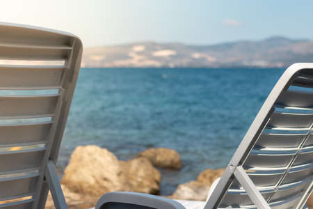 View of two white foldable benches near the sea, concept of vacation and summer. Bright blue water in the backgroundの写真素材