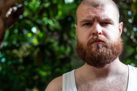 Bearded bald man in an undershirt with hairy chest standing outside looking straight at the camera. Serious sad look, feeling vulnerable and exposedの写真素材