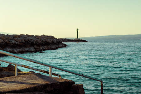 Ladder going down into the blue sea, rocky shore with a lighthouse in the distance. Moody morning in split,croatiaの写真素材