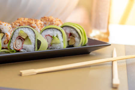 Homemade california and cucumber sushi rolls with surimi fake crab meat, avocado and fried sesame seeds. Served in two rows on a black plate at home. Chopsticks on table, bright sun background glareの写真素材