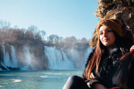 Attractive brunette posing on a wall looking into the distance. Kravice waterfall in the distance , bosnia and herzegovina. Cold winter dayの写真素材