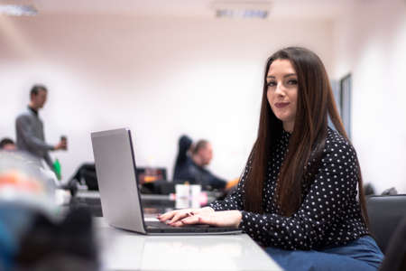 Young adult caucasian brunette woman sitting in an office environment looking at camera doing work. Multiple people in the background during a meeting.の写真素材