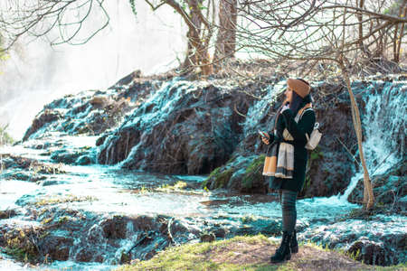 Girl traveller standing on a mossy patch of grass , cold river flowing in the background. Kravice waterfall in bosnia on a cold winter dayの写真素材
