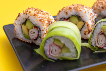Closeup view of a california sushi roll with sesame seeds and surimi in a sushi set on black plate and bright yellow backgroundの写真素材