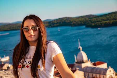 Portrait of an attractive brunette with a white shirt and sunglasses. Leaning on a wall , adriatic sea and the SIbenik cathedral in the distanceの写真素材