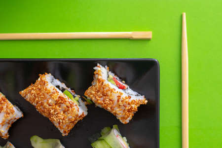 Top down view of a black square plate with multiple sushi rolls with sesame seeds and cucumber on green simple background with chopsticks paralel to the plateの写真素材