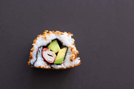 Closeup of a single piece of homemade not perfect sushi roll. Isolated on dark black background. Roll containing avocado, surimi, cucumber and fried sesame seedsの写真素材
