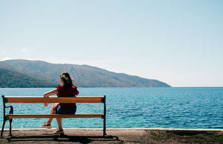 Unrecognisable woman seen from behind sitting on a bench on the seaside. Mountains and endless sea in the distance. Shore of Starigrad town Croatiaの写真素材