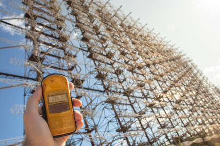 Hand holding a yellow dosimeter indicating radiation on the screen. Enormous Duga radar complex seen stretching to infinity in the distance. Danger of radiation in the exclusion zoneの写真素材
