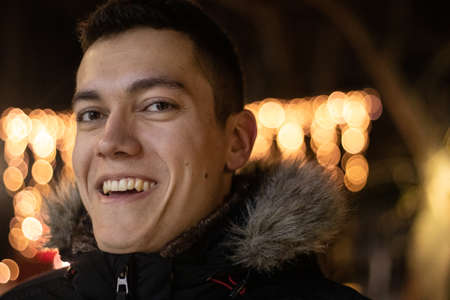 Headshot portrait of young man in winter, smiling and laughing at night with orange bokeh lights in...