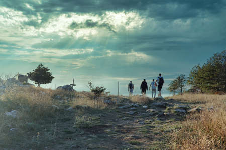 Four friends hiking together, reach the top of a small hill, stone path overgrown with grass leading to them. Dark mistical cloudy dayの写真素材