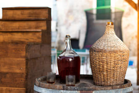 View of a glass bottle and a traditional wooden woven bottle holding several liters. Two empty glasses on the barrel. Small local meditarranean town, local domestic homemade wineの写真素材