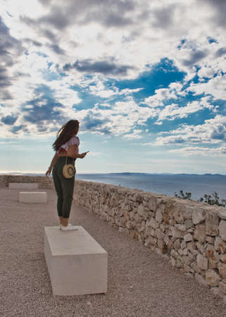 Brunette girl seen from behind standing on a stone bench on a windy day, hill observation point near...