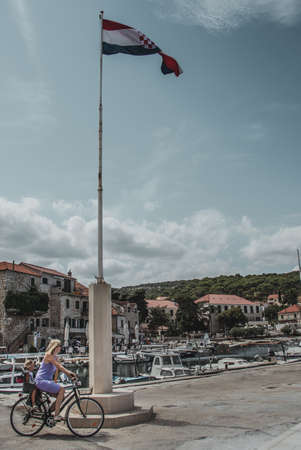Postira, Croatia August 2020 Mom cycling with a child around a tall pole with the Croatian flag on...