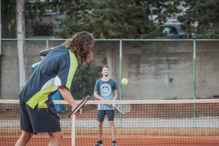 Man with longer hair seen from behind swining his racquet forehand to hit the ball, playing tennis outdoors with a friend. Ball seen blurred flying fast in the airの写真素材