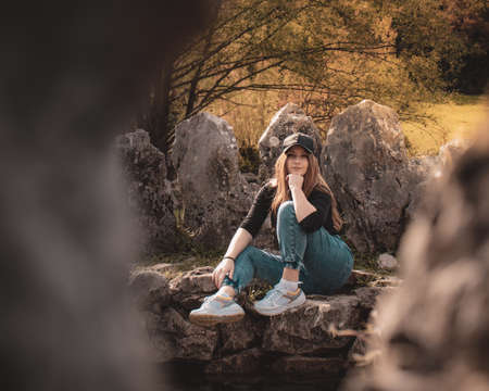 Zoomed full body portrait of an attractive brunette sitting on a well at the Rajcica wells natural landmark. Girl wearing jeans and a black cap, modern trending outfit on a warm brown autumn dayの写真素材