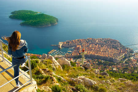 Attractive brunette traveller sitting on a railing above the city of Dubrovnik, viewpoint on Srd mountain. Looking at the ancient historic city surrounded by the blue adriatic seaの写真素材