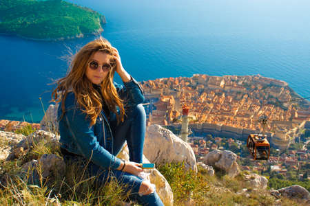 Attractive brunette sitting on the Srd hill above the city of Dubrovnik, observing the city in the distance. Wind blowing her hair above the ancient famous landmark town. Cablecar going downの写真素材
