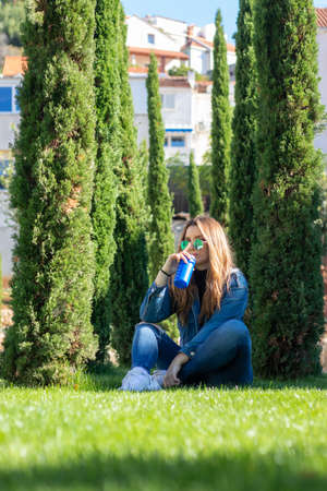 Vertical shot of an attractive brunette sitting in a grass field surrounded by trees, holding a blue sports bottle, relaxing and drinking water on a warm summer day. Concepts of health and relaxationの写真素材
