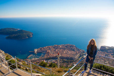 Brunette in blue jeans and top standing on the railing of Srd observation point above the city of Dubrovnik. Ancient city in the distance surrounded by the blue adriatic seaの写真素材