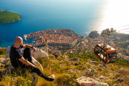 Wide shot from the top of the Srd mountain, man sitting on a rock observing the area around the city of Dubrovnik, orange cablecar descends to the city walls carrying tourist travellers in the summerの写真素材