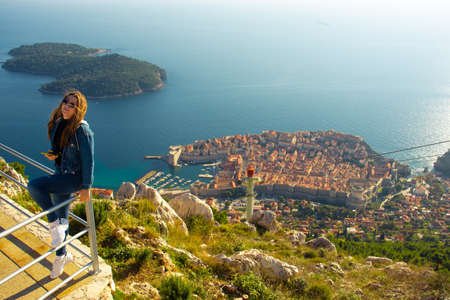 Attractive brunette traveller sitting on a railing above the city of Dubrovnik, viewpoint on Srd mountain. Looking at the ancient historic city surrounded by the blue adriatic seaの写真素材