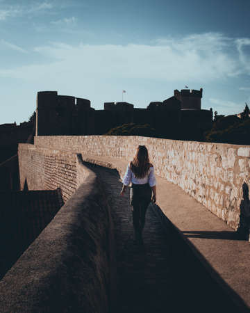 Vertical shot and rear view of an attractive girl walking along the city walls of Dubrovnik town. Dark moody edit with black and orange highlightsの写真素材