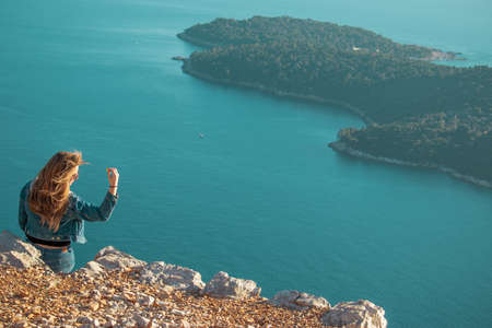 Rear view of an attractive brunette girl sitting on a stone posing, wind blowing her golden brown hair. Blue adriatic sea and the island of lokrum in the background. Travel concept with copyspace roomの写真素材