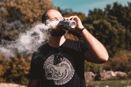 Split Croatia October 2020 Bald bearded man wearing a Game of thrones black shirt taking a sip of beer from a can, thick white vaping smoke leaving the can and forming a cloudのeditorial素材