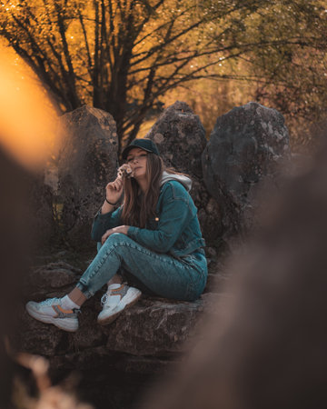 Portrait of an attractive brunette sitting in a stone well at the Rajcica wells landmark. Wearing jeans pants, jacket and a black cap, holding a bouquet of handpicked flowers on a cold autumn dayの写真素材