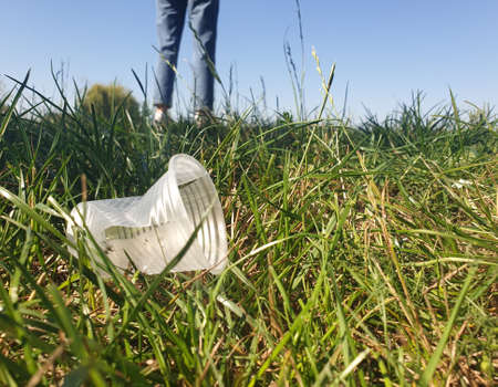 litter in a green grass: plastic cup on the ground, selective focus, blurred legs on background.