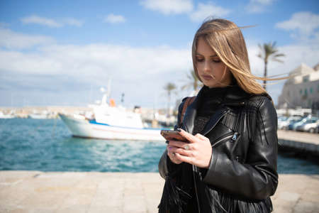 Beautiful blond-haired Italian woman whit clack jacket uses the phone on the port of a medieval seaside town in Molfetta Puglia in south Italyの写真素材