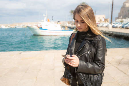 Beautiful blond-haired Italian woman whit clack jacket uses the phone on the port of a medieval seaside town in Molfetta Puglia in south Italyの写真素材