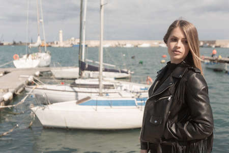 Young italian woman with long hair with leather black jacket looks in a marina with fishing boatsの写真素材