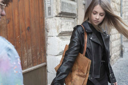 Beautiful young woman with straight blonde hair and black leather jacket and large bag in a medieval alley with her boyfriendの写真素材