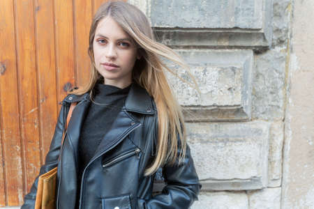Beautiful young italian woman with straight blonde hair and black leather jacket posing in a narrow medieval alley with a calm airの写真素材