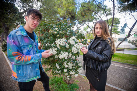 Outdoor, a young italian man and woman are couple in spring day in the park look over the hedge at the park flowers whit three and green whit flowerの写真素材